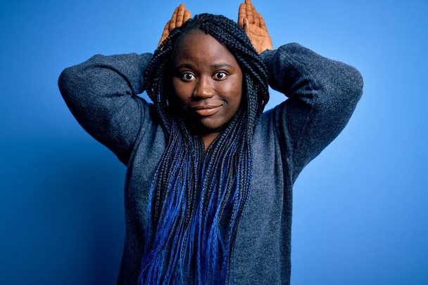 Afroamericano plus size donna con trecce che indossano maglione casual su sfondo blu Fare coniglietto orecchie gesto con le mani palme cercando cinico e scettico. Concetto di coniglio pasquale
. - Foto, immagini