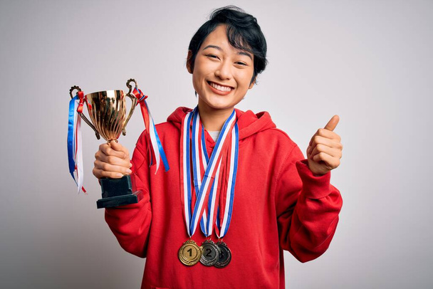 Young beautiful asian girl winner holding trophy wearing medals over white background happy with big smile doing ok sign, thumb up with fingers, excellent sign - Photo, Image