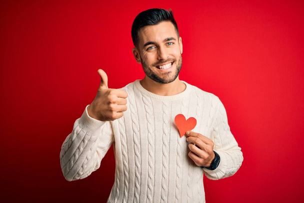 Kırmızı kalbi romantik ve sağlık sembolü olarak tutan yakışıklı genç adam gülümsüyor. Tamam işareti yapıyor, parmaklarıyla başparmağını kaldırıyor, mükemmel bir işaret. - Fotoğraf, Görsel