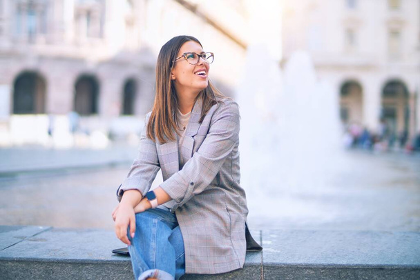 Młoda piękna kobieta uśmiechnięta szczęśliwa i pewna siebie. Siedząc z uśmiechem na twarzy na ulicy miasta - Zdjęcie, obraz