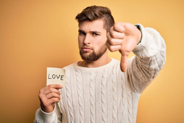Young blond romantic man with beard and blue eyes holding paper with love message with angry face, negative sign showing dislike with thumbs down, rejection concept - Photo, image