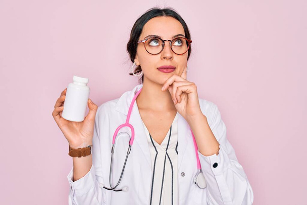 Young beautiful doctor woman with blue eyes wearing stethoscope holding medicine pills serious face thinking about question, very confused idea - Photo, Image