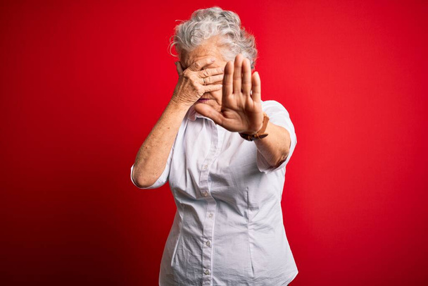 Senior kaunis nainen yllään tyylikäs paita seisoo yli eristetty punainen tausta peittää silmät kädet ja tekee stop ele surullinen ja pelko ilme. Hävettää ja negatiivinen käsite
. - Valokuva, kuva