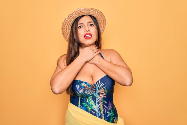 Young hispanic woman wearing summer hat and swimsuit over yellow background shouting and suffocate because painful strangle. Health problem. Asphyxiate and suicide concept. - Фото, зображення