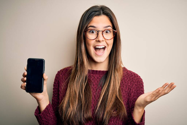 Young beautiful girl wearing glasses showing smartphone over isolated white background very happy and excited, winner expression celebrating victory screaming with big smile and raised hands - Photo, Image