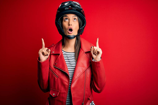 Young beautiful brunette motorcyclist woman wearing motorcycle helmet and red jacket amazed and surprised looking up and pointing with fingers and raised arms. - Фото, изображение