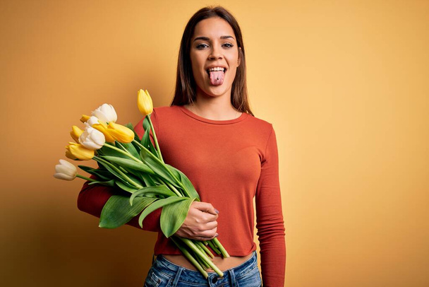 Nuori kaunis brunette nainen tilalla kimppu tulppaanit kukkia keltainen tausta törröttää kielen ulos onnellinen hauska ilme. Tunnekonsepti
. - Valokuva, kuva