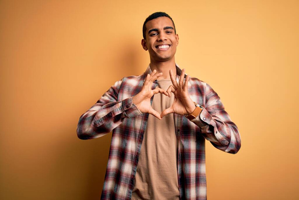 Mladý pohledný africký Američan nosí ležérní košili stojící nad žlutým pozadím s úsměvem v lásce dělá srdce symbol tvar s rukama. Romantický koncept. - Fotografie, Obrázek