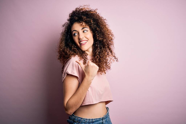 Young beautiful woman with curly hair and piercing wearing casual pink t-shirt smiling with happy face looking and pointing to the side with thumb up. - Фото, изображение