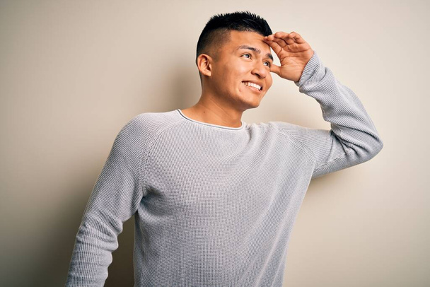 Young handsome latin man wearing casual sweater standing over isolated white background very happy and smiling looking far away with hand over head. Searching concept. - Photo, Image