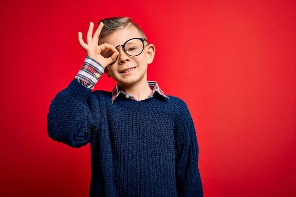 Young little caucasian kid with blue eyes standing wearing smart glasses over red background doing ok gesture with hand smiling, eye looking through fingers with happy face. - Photo, Image