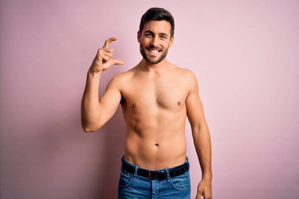 Young handsome strong man with beard shirtless standing over isolated pink background smiling and confident gesturing with hand doing small size sign with fingers looking and the camera. Measure concept. - Photo, Image
