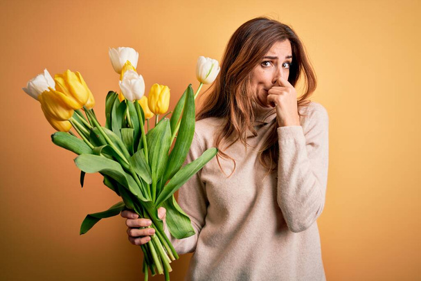 Young beautiful brunette woman holding bouquet of yellow tulips over isolated background smelling something stinky and disgusting, intolerable smell, holding breath with fingers on nose. Bad smell - Photo, Image