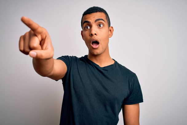 Молодий привабливий афроамериканець, одягнений у повсякденну футболку, що стоїть над білим фоном Очарований пальцем попереду, відкритий рот вражений вираз, щось на передньому плані - Фото, зображення