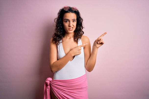 Schöne Frau mit lockigem Haar im Urlaub trägt weißen Badeanzug über rosa Hintergrund Zeigt besorgt und nervös mit beiden Händen, besorgt und überrascht Ausdruck - Foto, Bild