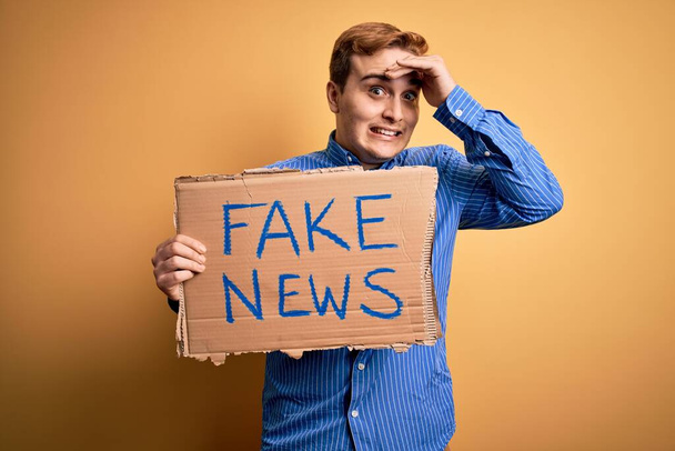 Junge hübsche rothaarige Mann mit Banner mit Fake-News-Nachricht über gelbem Hintergrund gestresst und frustriert mit Hand auf Kopf, überrascht und wütend Gesicht - Foto, Bild