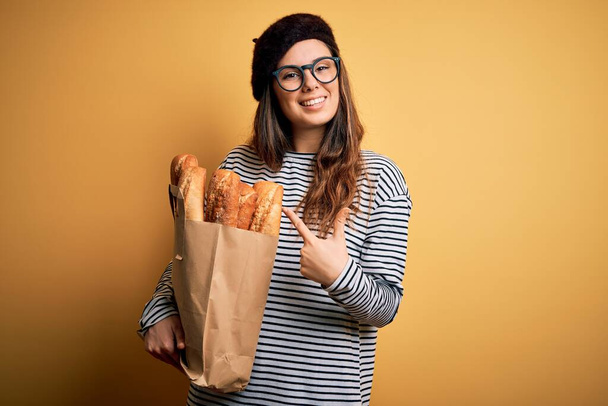 Young beautiful brunette french woman wearing beret holding paper bag with baguettes very happy pointing with hand and finger - Foto, immagini