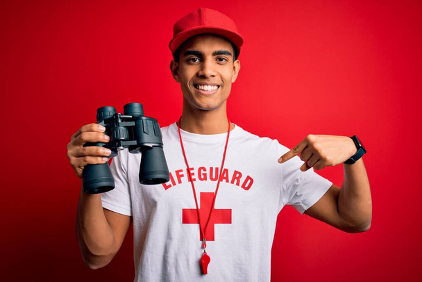 Young handsome african american lifeguard man wearing whistle using binoculars with surprise face pointing finger to himself - Foto, afbeelding