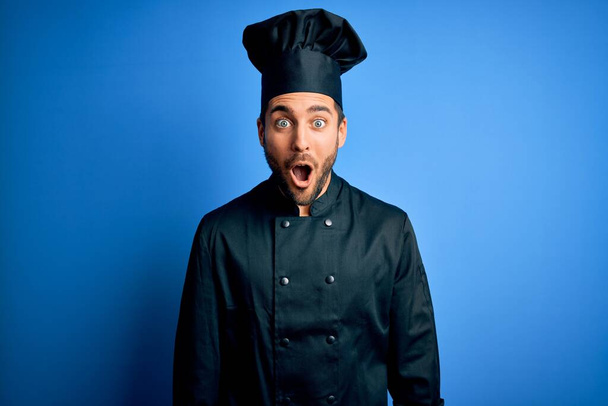 Joven hombre guapo chef con barba con uniforme de cocina y sombrero sobre fondo azul miedo y sorprendido con la expresión sorpresa, el miedo y la cara emocionada
. - Foto, imagen