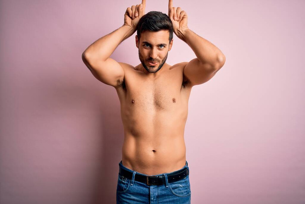 Young handsome strong man with beard shirtless standing over isolated pink background doing funny gesture with finger over head as bull horns - Foto, imagen