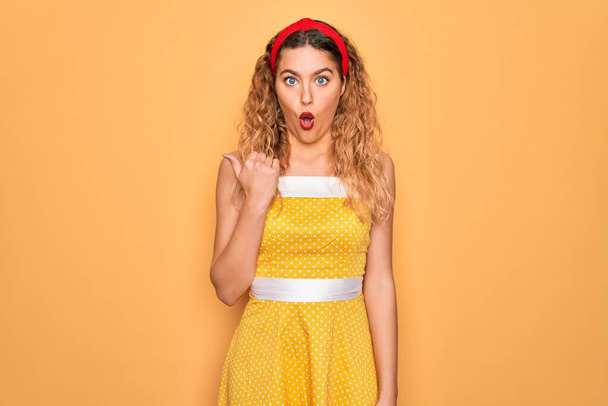 Mooie blonde pin-up vrouw met blauwe ogen dragen diadeem staande over gele achtergrond Verrast wijzend met de hand vinger naar de zijkant, open mond verbaasd uitdrukking. - Foto, afbeelding