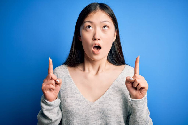 Молодая красивая азиатская женщина в обычном свитере, стоящем на синем изолированном фоне удивлена и удивлена, глядя вверх и указывая пальцами и поднятыми руками
. - Фото, изображение