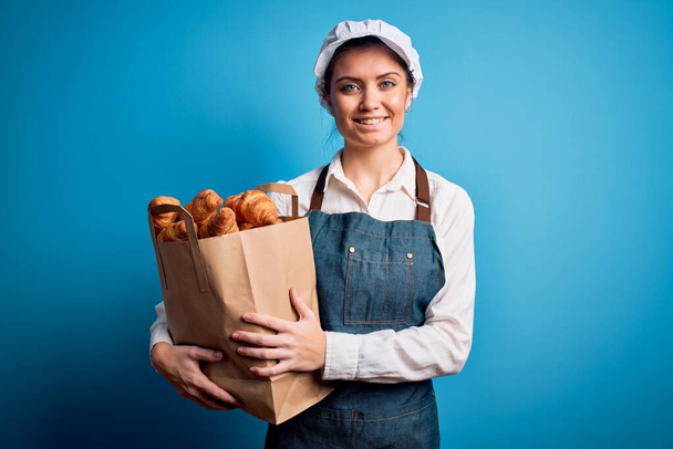 Joven hermosa panadera con ojos azules usando delantal sosteniendo bolsa de papel con cruasanes con una cara feliz de pie y sonriendo con una sonrisa confiada mostrando los dientes
 - Foto, imagen