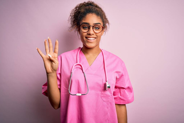 Niña enfermera afroamericana vistiendo uniforme médico y estetoscopio sobre fondo rosa mostrando y señalando con los dedos número cuatro mientras sonríe confiado y feliz
. - Foto, imagen