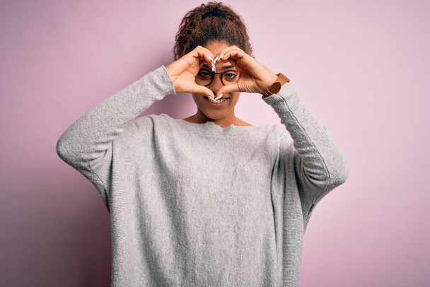 Young beautiful african american girl wearing sweater and glasses over pink background Doing heart shape with hand and fingers smiling looking through sign - Photo, Image