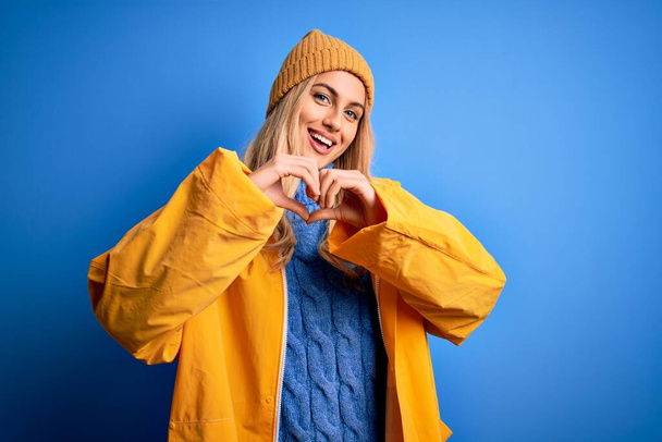 Jonge mooie blonde vrouw draagt regenjas voor regenachtig weer over blauwe achtergrond glimlachend in liefde met hart symbool en vorm met de handen. Romantisch concept. - Foto, afbeelding