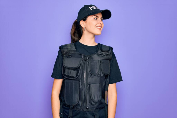 Молодая полицейская женщина в пуленепробиваемой форме на фиолетовом фоне смотрит в сторону с улыбкой на лице, естественным выражением лица. Смеясь уверенно
. - Фото, изображение
