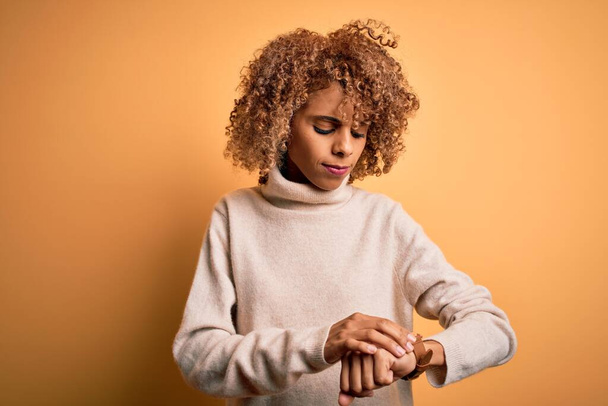Young beautiful african american woman wearing turtleneck sweater over yellow background Checking the time on wrist watch, relaxed and confident - Photo, Image