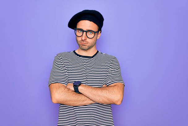 Handsome man with blue eyes wearing striped t-shirt and french beret over purple background skeptic and nervous, disapproving expression on face with crossed arms. Negative person. - Fotografie, Obrázek