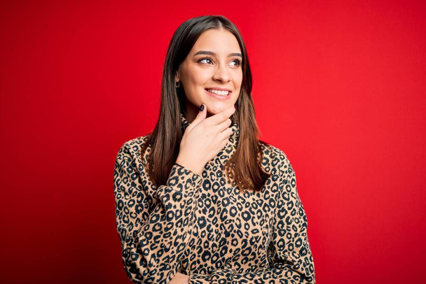 Young beautiful brunette woman wearing casual sweater standing over red background with hand on chin thinking about question, pensive expression. Smiling with thoughtful face. Doubt concept. - 写真・画像