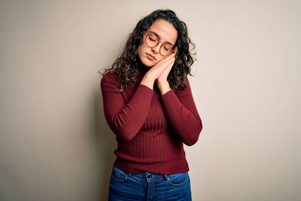 Beautiful woman with curly hair wearing casual sweater and glasses over white background sleeping tired dreaming and posing with hands together while smiling with closed eyes. - Photo, Image