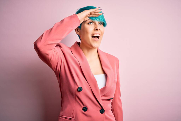 Mladá krásná podnikatelka s modrými módními vlasy nosí bundu přes růžové pozadí velmi šťastný a usmívá se daleko pryč s rukou nad hlavou. Vyhledávací koncept. - Fotografie, Obrázek