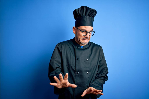 Young handsome chef man wearing cooker uniform and hat over isolated blue background disgusted expression, displeased and fearful doing disgust face because aversion reaction. - Photo, Image