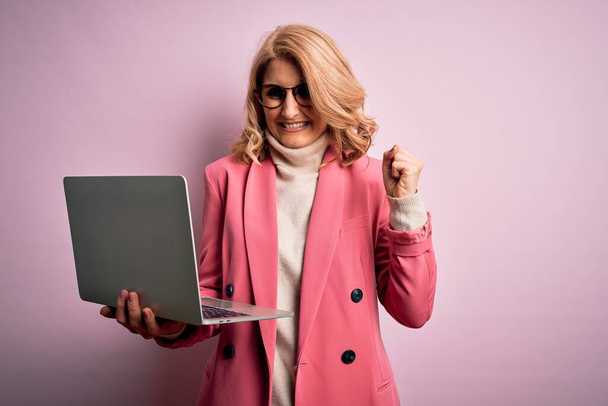Schöne blonde Geschäftsfrau mittleren Alters arbeitet mit Laptop vor rosa Hintergrund schreit stolz und feiert Sieg und Erfolg sehr aufgeregt, jubelnde Emotion - Foto, Bild