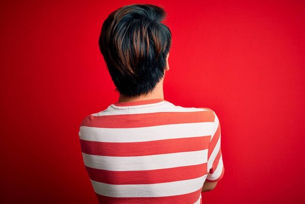 Nuori komea kiinalainen mies päällään rento raidallinen t-paita seisoo yli punainen tausta seisoo taaksepäin katsoen pois ristissä kädet
 - Valokuva, kuva