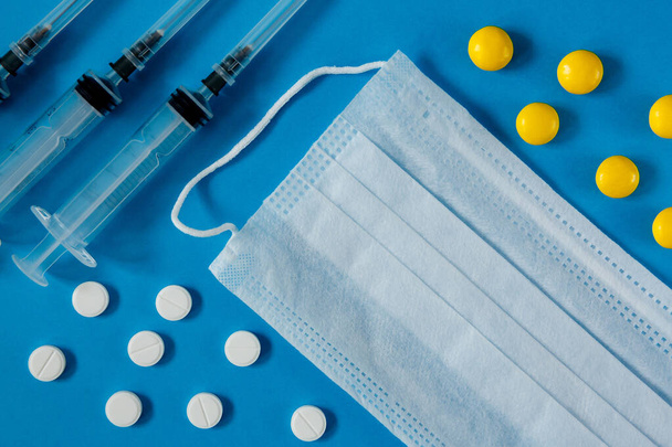 medical mask on the face, syringes and white and yellow pills, on a blue background from above - Photo, image