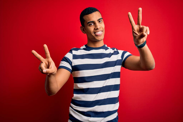 Handsome african american man wearing casual striped t-shirt standing over red background smiling with tongue out showing fingers of both hands doing victory sign. Number two. - Foto, immagini