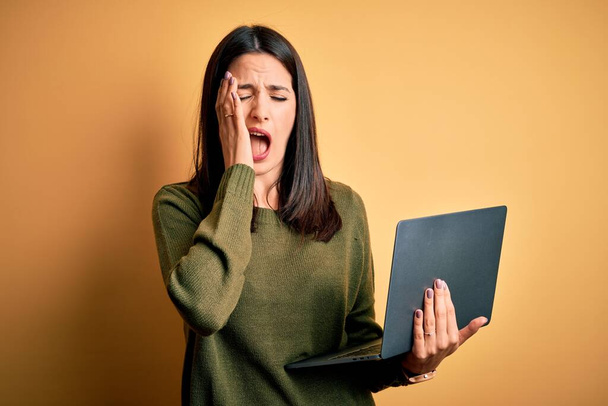 Young brunette woman with blue eyes working using computer laptop over yellow background Yawning tired covering half face, eye and mouth with hand. Face hurts in pain. - Photo, Image