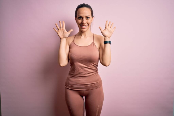 Молодая блондинка фитнес-женщина в спортивной одежде тренировки на розовом изолированном фоне показывая и показывая пальцами номер десять улыбаясь уверенно и счастливо
. - Фото, изображение