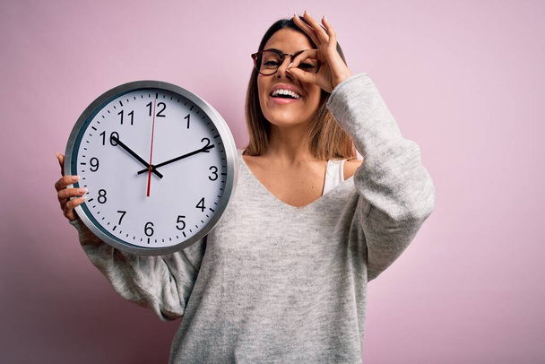 Young beautiful brunette woman wearing glasses doing countdown using big clock with happy face smiling doing ok sign with hand on eye looking through fingers - Foto, immagini