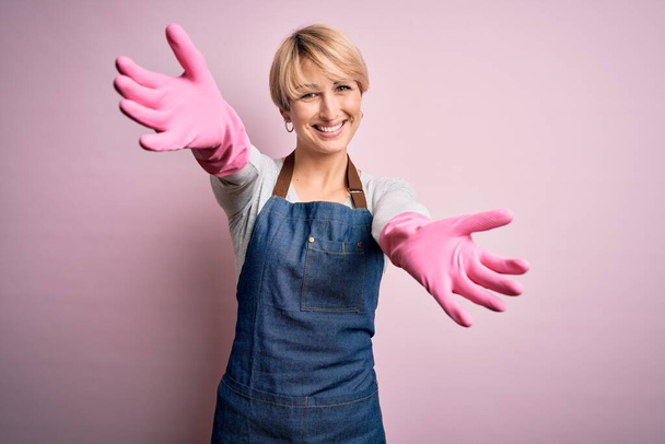 Junge blonde Putzfrau mit kurzen Haaren, Schürze und Handschuhen über rosa Hintergrund und lächelnd mit offenen Armen zur Umarmung in die Kamera. Fröhlicher Ausdruck des Glücks. - Foto, Bild