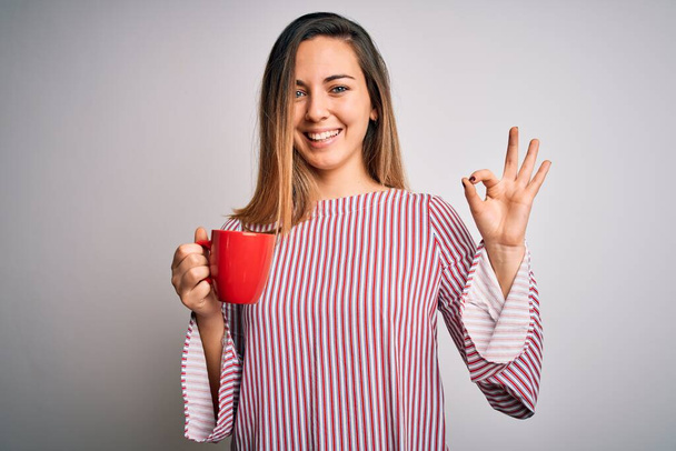 Beautiful blonde woman with blue eyes drinking red mug of coffee over white background doing ok sign with fingers, excellent symbol - Photo, Image