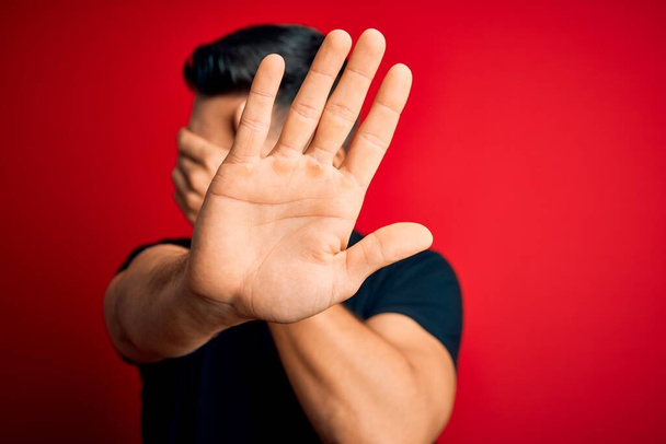 Jonge knappe man draagt casual zwarte t-shirt staan over geïsoleerde rode achtergrond bedekken ogen met handen en doen stop gebaar met droevige en angst uitdrukking. Beschaamd en negatief concept. - Foto, afbeelding