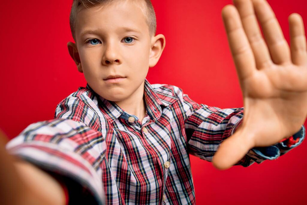 zbliżenie się młody mały kaukaski dzieciak z niebieskimi oczami biorąc selfie zdjęcie nad czerwonym tle z otwartą ręką robi znak stop z poważnym i pewnym siebie wyrazem twarzy, obrona gest - Zdjęcie, obraz