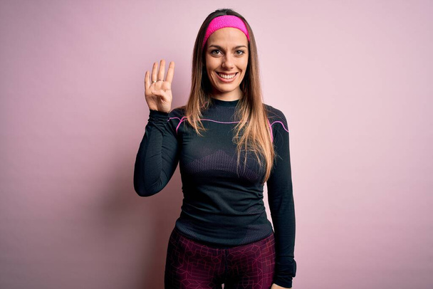Młoda blondynka fitness kobieta noszenie sportowe ubrania treningowe na odosobnionym tle pokazując i wskazując palcem numer cztery uśmiechając się pewny siebie i szczęśliwy. - Zdjęcie, obraz