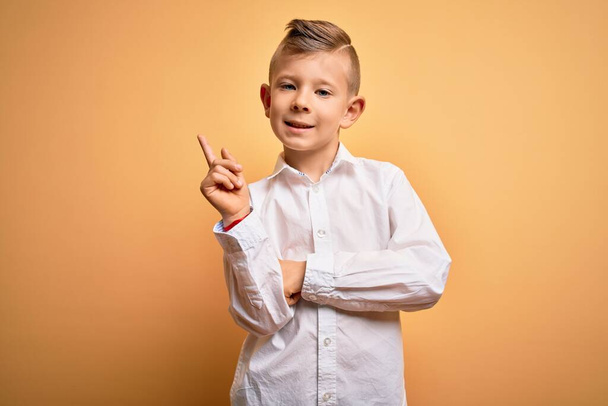 Junge kleine kaukasische Kinder mit blauen Augen tragen elegantes weißes Hemd über gelbem Hintergrund mit einem breiten Lächeln im Gesicht und zeigen mit der Hand und dem Finger zur Seite in die Kamera. - Foto, Bild
